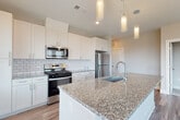 This thoughtfully designed single-floor residence combines contemporary finishes with a clean, functional layout. The open-concept kitchen is a standout feature, showcasing crisp white shaker cabinetry, a classic subway tile backsplash, speckled granite countertops, and a full suite of stainless steel appliances including a gas range, microwave, and refrigerator. A generously sized center island with a built-in sink and elegant pendant lighting above adds both practicality and visual appeal. The kitchen opens to a light-filled living area with wood-look flooring and easy access to an exterior door, enhancing the sense of openness throughout. The bedrooms are finished with plush carpeting and feature large windows fitted with blinds that usher in natural light and offer views of the surrounding neighborhood. The primary bathroom is well-appointed with a glass-enclosed walk-in shower clad in subway tile, a granite-topped vanity, wood-look flooring, and direct access to a dedicated walk-in closet. A secondary bathroom mirrors the refined aesthetic with a full subway-tiled tub and shower combination, a granite vanity, and matching wood-look floors. Neutral tones, clean lines, and consistent finishes throughout create a cohesive and inviting atmosphere across the entire space.