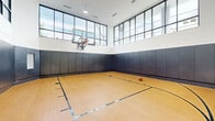 Located at 5711 John F Kennedy Blvd in North Bergen, NJ, this indoor basketball court is a standout athletic facility designed for serious play and active recreation. The court features a regulation hardwood-style floor with clearly marked boundary lines, three-point arcs, and key areas, providing an authentic playing experience. A professional-grade glass backboard and hoop are mounted at regulation height, underscoring the court's commitment to quality.

The space impresses with its soaring ceiling height, which accommodates full-court play with ease. Expansive floor-to-ceiling steel-framed windows line the upper walls on multiple sides, flooding the interior with generous natural light and offering open sky views that enhance the sense of airiness throughout the day. The lower walls are fully lined with padded safety panels in a sleek dark gray finish, offering both player protection and a modern, polished aesthetic.

An interior viewing window and an elevated glazed observation area allow spectators to watch activity from adjacent spaces. A bench along the sideline provides seating for players at rest. The overall design blends functionality with contemporary architecture, making this court a premier amenity well-suited for recreational leagues, personal training, and competitive play alike.