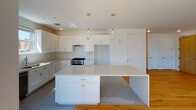 This single-floor residence showcases a clean, contemporary design throughout. The open-concept living and dining area is immediately striking, with dramatically tall ceilings and an abundance of natural light streaming through oversized windows that frame urban views of the surrounding neighborhood. Warm, light-toned hardwood flooring runs continuously through the main living spaces, complementing the crisp white walls and creating an inviting, airy atmosphere.

The kitchen is well-appointed with white shaker-style cabinetry, a stainless steel range, and a dishwasher. A generously sized central island with a white quartz countertop provides ample workspace and casual seating. Two glass pendant lights hang above the island, adding a refined touch. The kitchen floor features subtle gray subway-style tile that transitions smoothly into the hardwood of the living area.

The bedrooms continue the bright, modern aesthetic, each featuring hardwood floors, ceiling fans, and windows with outdoor views. The bathrooms are finished with hexagonal mosaic tile flooring, white subway tile surrounds in the shower and tub areas, white vanities, and chrome fixtures, delivering a timeless and polished look throughout.