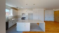 This single-floor residence showcases a clean, contemporary design throughout. The open-concept living and dining area is immediately striking, with dramatically tall ceilings and an abundance of natural light streaming through oversized windows that frame urban views of the surrounding neighborhood. Warm, light-toned hardwood flooring runs continuously through the main living spaces, complementing the crisp white walls and creating an inviting, airy atmosphere.

The kitchen is well-appointed with white shaker-style cabinetry, a stainless steel range, and a dishwasher. A generously sized central island with a white quartz countertop provides ample workspace and casual seating. Two glass pendant lights hang above the island, adding a refined touch. The kitchen floor features subtle gray subway-style tile that transitions smoothly into the hardwood of the living area.

The bedrooms continue the bright, modern aesthetic, each featuring hardwood floors, ceiling fans, and windows with outdoor views. The bathrooms are finished with hexagonal mosaic tile flooring, white subway tile surrounds in the shower and tub areas, white vanities, and chrome fixtures, delivering a timeless and polished look throughout.