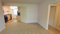 This single-floor space features a clean, understated design with neutral cream-toned walls and wall-to-wall beige carpeting in the main living area, creating a warm and versatile backdrop suitable for various furniture arrangements. The living space is open and uncluttered, with white baseboards providing a subtle contrast to the soft carpet and walls.

An open passthrough connects the living area to the kitchen, which is visible in the background and features light wood cabinetry paired with dark, contrasting countertops and a dark tile or stone backsplash. A window in the kitchen allows natural light to filter into the space, brightening both the kitchen and the adjacent living area. The kitchen appears to offer adequate counter and storage space for everyday use.

The overall layout promotes an easy flow between the living and kitchen areas, making the space practical for daily living. The simple, neutral finishes throughout provide a blank canvas that can be easily personalized with furnishings and décor.