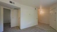 This single-floor space presents a straightforward, functional layout characterized by clean white walls and neutral carpet flooring that runs throughout the interior. The main living area is open and uncluttered, offering a versatile foundation for various furniture arrangements. A central wall division separates distinct zones within the space, with a door providing access to an adjoining room or hallway. Visible HVAC vents in both the ceiling and walls indicate a ducted climate control system serving the space.

To the right, a warm-toned area is illuminated by a modest chandelier, adding a decorative touch and a sense of warmth to an otherwise understated interior. This section appears to function as a transitional or dining area, benefiting from the ambient glow of the light fixture overhead. To the left, a hallway leads to additional rooms, suggesting further functional spaces beyond what is immediately visible.

The overall ambiance is neutral and move-in ready, with a blank-canvas aesthetic defined by cream-colored walls, consistent flooring, and practical finishes throughout. The layout supports easy customization to suit a variety of uses.