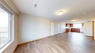This single-floor space offers a clean, open layout that connects the main living area to the kitchen in a practical, flowing arrangement. The living area is generously proportioned, featuring neutral white walls, warm wood-toned baseboards, and durable wood-look laminate flooring that extends throughout the space, lending a cohesive and modern aesthetic. The flat ceiling is finished cleanly and includes a central flush-mount light fixture along with a centrally placed HVAC vent, ensuring efficient climate control.

Two interior doors are visible along the back wall of the living area, likely leading to additional rooms or utility spaces, adding to the functional layout of the home. The open passage leads directly into the kitchen, which features rich, warm-toned wood cabinetry, a stainless steel appliance suite, and a sink positioned beneath additional upper cabinetry. The kitchen benefits from its own overhead lighting, keeping the space bright and practical. Overall, the space presents a straightforward, well-maintained interior with a neutral palette, easy-to-clean flooring, and an open feel suited to everyday living.