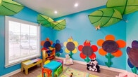 This cheerful, single-floor space is thoughtfully designed as a dedicated children's play area, bursting with color and creativity at every turn. The standout feature is the vivid sky-blue walls adorned with large, hand-crafted sunflower murals in bold shades of red, orange, yellow, and purple — each with a chalkboard center that invites interactive engagement. Oversized green leaf canopies hang from the ceiling alongside recessed lighting, creating an immersive garden-in-the-sky atmosphere. Decorative butterflies accent the walls, adding whimsical detail throughout. The room is well-lit by natural light streaming through windows with blinds, complemented by recessed ceiling fixtures. Light wood-look flooring runs throughout, providing a durable and easy-to-maintain surface. Child-sized colorful tables and chairs offer a dedicated activity area, while a multi-bin toy organizer keeps supplies neatly accessible. A stepped white cube storage unit lines one wall, topped with plush toys and building blocks, with a wall-mounted flat-screen TV positioned above. Plush animal seating adds a playful, cozy touch. The space also includes a hallway corridor with ADA-compliant dual water fountains and clearly marked restroom facilities, ensuring the space is fully accessible and functional for families and caregivers.