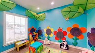 This cheerful, single-floor space is thoughtfully designed as a dedicated children's play area, bursting with color and creativity at every turn. The standout feature is the vivid sky-blue walls adorned with large, hand-crafted sunflower murals in bold shades of red, orange, yellow, and purple — each with a chalkboard center that invites interactive engagement. Oversized green leaf canopies hang from the ceiling alongside recessed lighting, creating an immersive garden-in-the-sky atmosphere. Decorative butterflies accent the walls, adding whimsical detail throughout. The room is well-lit by natural light streaming through windows with blinds, complemented by recessed ceiling fixtures. Light wood-look flooring runs throughout, providing a durable and easy-to-maintain surface. Child-sized colorful tables and chairs offer a dedicated activity area, while a multi-bin toy organizer keeps supplies neatly accessible. A stepped white cube storage unit lines one wall, topped with plush toys and building blocks, with a wall-mounted flat-screen TV positioned above. Plush animal seating adds a playful, cozy touch. The space also includes a hallway corridor with ADA-compliant dual water fountains and clearly marked restroom facilities, ensuring the space is fully accessible and functional for families and caregivers.