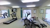 This single-floor fitness room provides a practical and functional space for exercise, featuring a selection of cardio and strength training equipment. The room is outfitted with a treadmill, an elliptical trainer, a stationary bike, and an air resistance exercise machine, offering a well-rounded range of workout options. A flat-screen TV is wall-mounted near the treadmill, allowing users to stay entertained during their workouts. A workout reference chart is posted on the wall, serving as a helpful guide for exercise routines. The room is illuminated by a pair of large flush-mounted fluorescent ceiling fixtures that provide bright, even lighting throughout the space. Vertical blinds cover a window along one wall, allowing for adjustable natural light. The flooring consists of low-pile carpet, offering a comfortable surface underfoot. The walls are painted in a neutral cream tone, accented by a teal-colored vertical stripe, adding a subtle decorative touch. The overall layout is organized and efficient, making practical use of the available space to accommodate multiple users and pieces of equipment simultaneously.