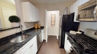 This single-story, one-bedroom, one-bathroom residence offers a thoughtfully designed layout that maximizes everyday functionality. The kitchen serves as a standout feature, designed in a classic galley style that efficiently utilizes the available space. White shaker-style cabinetry lines both sides of the kitchen, providing ample storage from floor to ceiling. Dark granite countertops offer a striking contrast against the white cabinetry, lending the space a polished, contemporary aesthetic. Black appliances, including a refrigerator and a gas range, complement the countertops, while a stainless steel sink and dishwasher add a modern touch. The kitchen flows toward an interior doorway that connects to additional areas of the residence, suggesting a practical and cohesive floor plan. Wood-style flooring runs throughout the kitchen, contributing warmth and continuity to the space. The neutral wall tones create a clean, versatile backdrop that pairs well with the kitchen's design elements. Overall, this residence combines classic design choices with functional details, making it a comfortable and well-equipped single-floor living space.