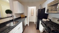 This single-story, one-bedroom, one-bathroom residence offers a thoughtfully designed layout that maximizes everyday functionality. The kitchen serves as a standout feature, designed in a classic galley style that efficiently utilizes the available space. White shaker-style cabinetry lines both sides of the kitchen, providing ample storage from floor to ceiling. Dark granite countertops offer a striking contrast against the white cabinetry, lending the space a polished, contemporary aesthetic. Black appliances, including a refrigerator and a gas range, complement the countertops, while a stainless steel sink and dishwasher add a modern touch. The kitchen flows toward an interior doorway that connects to additional areas of the residence, suggesting a practical and cohesive floor plan. Wood-style flooring runs throughout the kitchen, contributing warmth and continuity to the space. The neutral wall tones create a clean, versatile backdrop that pairs well with the kitchen's design elements. Overall, this residence combines classic design choices with functional details, making it a comfortable and well-equipped single-floor living space.