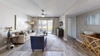 This single-floor residence offers a thoughtfully designed layout that balances comfort and functionality throughout its interconnected living spaces. The living room serves as the heart of the home, featuring wood-look flooring, a ceiling fan, and a sliding glass door that opens to an outdoor patio, flooding the space with natural light and providing pleasant exterior views. The adjacent dining area accommodates a round table with upholstered seating and flows seamlessly into the kitchen, which is equipped with white shaker-style cabinetry, quartz countertops, a white subway tile backsplash, and black appliances including a range and dishwasher. The bedroom is generously proportioned, featuring large sliding glass doors with vertical blinds and horizontal shades that allow abundant natural light, complemented by wood-look flooring and a spacious walk-in closet with wire shelving for organized storage. The bathrooms include a tub-shower combination, with one featuring white tile surround and gold fixtures. Throughout the unit, a neutral gray color palette, consistent wood-look flooring, and clean white trim create a cohesive, modern aesthetic.