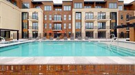 Located at 6121 S Highland Dr in Holladay, Utah, this thoughtfully designed outdoor amenity space offers a comprehensive resort-style experience within a contemporary mixed-use development. The centerpiece is a generously sized swimming pool with sparkling turquoise water, framed by a brick coping detail and lined with rows of sleek chaise lounges — ideal for relaxing and sunbathing. Adjacent to the pool, a tiled terrace provides additional seating with teak wood dining sets shaded by vibrant orange umbrellas, accompanied by overhead radiant heaters for year-round comfort.

The surrounding multi-story building showcases a modern architectural palette, blending warm brick, horizontal wood cladding, and light stucco with large-format windows and private balconies. A spacious open-air courtyard connects to an interior space through floor-to-ceiling glass walls, allowing seamless indoor-outdoor flow. String lights and pergola structures with horizontal slatted roofs define intimate lounge areas furnished with contemporary sectional seating throughout the terrace.

An upper-level rooftop deck extends the outdoor living space further, featuring large-format porcelain tile flooring, additional pergola cabanas with cushioned seating, and open views of the surrounding Holladay neighborhood. The cohesive design and high-quality finishes throughout create a polished, inviting atmosphere suited for both relaxation and social gatherings.