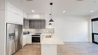 This single-floor residence showcases a clean, modern aesthetic with an open-concept layout that seamlessly connects the kitchen and living areas. The kitchen is a standout feature, equipped with a large island topped with a bright white quartz-style countertop and an integrated sink, complemented by a dual-tone cabinet design pairing crisp white upper cabinets with rich wood-grain lower cabinets. A textured tile backsplash adds depth, while stainless steel appliances—including a refrigerator with ice maker, range, and built-in microwave—provide a sleek, functional finish. Stylish dark bronze pendant lights hang above the island, anchoring the space with a contemporary touch. Recessed lighting and light wood-tone flooring run throughout, lending warmth and continuity across the open living area, which benefits from generous natural light through large glass doors that open to an exterior balcony. The bedroom offers a calm, neutral retreat, while the bathroom features a walk-in shower with curtain surround, a white vanity with ample drawer storage, and coordinating matte black hardware. A generously sized walk-in closet outfitted with wire shelving and hanging rods provides substantial storage. The overall design emphasizes minimalism, functionality, and contemporary style.