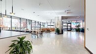 This contemporary single-floor amenity space is defined by its open-plan layout, polished concrete flooring, and an abundance of natural light streaming through floor-to-ceiling black-framed glass walls that offer unobstructed views of the surrounding cityscape. The main lounge area is anchored by a generously sized tan leather sectional sofa, a linear gas fireplace set into a stone surround, and a large-format wall-mounted display above it, creating a refined yet comfortable gathering space. Adjacent to the lounge, a communal dining area provides ample seating for groups, complemented by lush indoor greenery that adds warmth and texture throughout. The fully equipped kitchen features sleek dark cabinetry, integrated appliances, stone countertops, a substantial island with bar seating, and pendant lighting — all framed by city views through the glass facade. A dedicated game room includes a billiards table set against a rich wood-paneled accent wall, along with a mounted display and bar-height seating. A glass-railed staircase connects to a lower level, offering additional spatial depth. The overall design blends industrial and contemporary elements — polished surfaces, bold structural columns, curated artwork, and abundant plantings — to create a vibrant, multifunctional community environment.