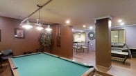 This single-floor space is thoughtfully designed for entertainment and relaxation. The centerpiece of the main area is a full-sized pool table, positioned beneath a stylish multi-pendant chandelier that casts warm, focused light over the playing surface. Rich, deep brown walls create a cozy and immersive atmosphere, complemented by recessed ceiling lighting throughout. A decorative artificial tree adds a touch of organic warmth to the corner of the room, while a mounted cue rack keeps billiards equipment neatly organized and within easy reach.

An adjoining area transitions seamlessly from the game room, featuring a casual dining set with wrought-iron style chairs gathered around a glass-top table. This secondary space is finished in a contrasting lighter tone, giving it a distinct yet cohesive feel. A window with vertical blinds allows for natural light, and a decorative circular wall accent adds a artistic focal point to the room.

Together, the spaces offer a well-balanced environment suited for both active recreation and casual dining, making this layout an ideal setting for social entertaining. The combination of warm tones, ambient lighting, and purposeful furnishings gives the space a welcoming and functional character.
