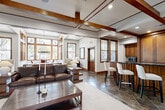 Located at 5045 Crenshaw Rd in Pasadena, TX, this single-floor community clubhouse offers a thoughtfully designed gathering space defined by warm craftsman-style architecture. Slate tile flooring runs throughout, complemented by rich wood trim, paneling, and window surrounds that lend a cohesive, upscale aesthetic.

The spacious entry foyer welcomes visitors with double wood-and-glass doors, large sidelight windows that flood the space with natural light, and open sightlines into adjoining areas. A dedicated office nook is accessible directly from the foyer.

The main lounge is a standout, featuring an exposed wood beam ceiling with coffered detailing, recessed lighting, and a fireplace flanked by built-in wood shelving and a wall-mounted TV. A full-size pool table anchors the game area, while a leather sofa and wood coffee table define a comfortable seating zone nearby.

The wet bar area is outfitted with dark granite countertops, warm wood cabinetry, a built-in microwave, and high-top bar seating — ideal for casual entertaining.

The dining area is bathed in natural light from a dramatic wraparound window arrangement, offering views of the surrounding landscape. A round wood dining table beneath a striking wrought-iron chandelier completes the refined yet inviting atmosphere of this versatile community space.