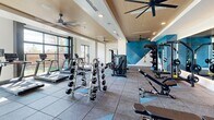 Located at 414 Front St in Santa Cruz, CA, this fitness center offers a thoughtfully designed exercise space that combines functionality with modern aesthetics. The single-floor layout provides a generous, open floor plan that comfortably accommodates a full range of workout equipment without feeling crowded.

The space features durable rubber flooring with blue geometric accent tiles, providing both safety and visual interest throughout. Overhead, warm wood-paneled ceiling insets are complemented by recessed lighting and multiple ceiling fans, contributing to a well-ventilated and inviting atmosphere. Large floor-to-ceiling windows along the perimeter flood the room with natural light and offer views of the surrounding outdoor environment.

The exercise room is comprehensively equipped with a robust selection of True-brand treadmills arranged along the windowed wall, multiple cable and strength machines, dumbbell racks with a full range of free weights, adjustable benches, and TRX suspension training equipment. A full-length wall mirror expands the perceived depth of the space while allowing users to monitor their form. A bold teal geometric accent wall adds a striking design element and visual energy to the room. Built-in shelving provides convenient storage for accessories. The overall ambiance is modern, clean, and energizing — well-suited for a comprehensive fitness experience.