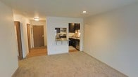 This single-floor residential unit offers a functional and well-appointed layout designed for comfortable everyday living. The open living area is finished with plush carpet flooring, creating a warm and inviting atmosphere, while recessed lighting and a flush-mount ceiling fixture provide ample illumination throughout the space. A solid entry door with a security peephole opens into a transitional area featuring hardwood-style flooring, offering a clean separation between the entry and the main living space. The kitchen is efficiently designed and tucked into a defined alcove, featuring dark espresso cabinetry that contrasts elegantly against the light-colored walls. Stainless steel appliances, including a refrigerator, microwave, and range, lend a modern touch, while a single-basin sink with a window above adds both practicality and natural light. A built-in breakfast bar with a countertop overhang bridges the kitchen and living area, making it ideal for casual dining or entertaining. The overall aesthetic is clean and contemporary, with neutral tones throughout that offer a versatile backdrop for a variety of interior styles. The thoughtful layout maximizes the use of available space, making this unit both practical and visually appealing.