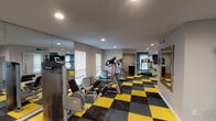 Located at 2121 Tannehill in Houston's 77008 zip code, this single-floor fitness center is a well-appointed exercise facility designed to support a comprehensive workout experience. The main workout area is immediately distinguished by its bold yellow and charcoal checkered carpet tile flooring, which lends the space a dynamic, energetic aesthetic. Recessed lighting throughout ensures the room is consistently and evenly illuminated, complementing the natural light admitted through multiple windows fitted with roller shades.

The facility is equipped with a robust selection of cardio machines, including treadmills, ellipticals, and spin bikes, as well as strength-training equipment and resistance machines. Large floor-to-ceiling mirrors line the walls, visually expanding the space and allowing users to monitor their form during workouts. Wall-mounted televisions provide entertainment options during exercise sessions. Stability balls and additional accessories are also available for functional training.

Adjacent to the main workout floor is a dedicated kitchenette area, finished with rich dark wood cabinetry, granite-style countertops, a tile backsplash, hardwood flooring, and stainless steel appliances — offering a convenient space for post-workout refreshment or preparation. Together, these areas create a functional, modern, and well-rounded fitness facility.