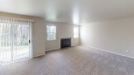 Located at 7688 Vineyards Ln NE in Bremerton, Washington, this single-floor unit offers a practical and comfortable layout throughout. The spacious living room serves as the heart of the home, featuring plush carpeting, a classic fireplace flanked by two windows that invite natural light, and a sliding glass door leading to an outdoor area. The open sight line from the kitchen counter into the living space creates a connected, airy feel. The galley-style kitchen is well-equipped with white cabinetry offering ample storage, laminate countertops, a black electric range, dishwasher, and a full-size refrigerator, all set on durable stone-tile-style flooring. A carpeted hallway connects the living areas to the private spaces, including bedrooms with windows overlooking the outdoors and closet access. The full bathroom features a vanity with a single sink, white cabinetry, a toilet, and a tub/shower combination with a curtain rod, finished with the same tile-style flooring found in the kitchen. Neutral tones, textured walls, and consistent finishes throughout give the unit a clean, cohesive aesthetic suited for comfortable everyday living.