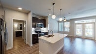 This contemporary single-floor residence showcases a thoughtfully designed open-concept layout that seamlessly connects the kitchen, dining, and living areas. The kitchen is a standout feature, anchored by a large center island with a white quartz countertop and an undermount sink with a brushed gold faucet. Rich dark espresso cabinetry extends to the ceiling, providing ample storage, while stainless steel appliances — including a French door refrigerator with ice dispenser, electric range, over-the-range microwave, and dishwasher — complete the chef-ready space. Globe pendant lights hang above the island, adding a modern, polished aesthetic. The living area is bright and open, featuring warm wood-look flooring that flows throughout the space, a ceiling fan, and a wall of windows alongside French doors that open to a private balcony, allowing generous natural light and outdoor views. The bathroom continues the refined design language with dark cabinetry, a white quartz vanity, framed mirrors, sconce lighting, and a tiled tub-shower combination. An in-unit washer and dryer are also visible, adding everyday convenience. Neutral wall tones and clean architectural lines give the entire space a cohesive, contemporary feel.