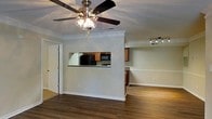 This single-floor residence offers a thoughtfully designed open-concept layout that seamlessly connects the living and kitchen areas. The living space features rich hardwood-style flooring, elegant crown molding, and a ceiling fan with integrated lighting, contributing to a comfortable and polished atmosphere. A distinctive pass-through window in the half-wall connects the living area to the kitchen, creating an open and airy feel while maintaining a sense of defined space. The kitchen is equipped with wooden cabinetry in a warm honey tone, dark granite or stone countertops, and black appliances including a refrigerator and microwave. Track lighting above the kitchen area provides focused illumination for both functionality and ambiance. An adjacent dining or secondary living area is visible beyond the kitchen, also featuring track lighting and the same consistent crown molding detail found throughout the home. Crisp white walls and white baseboards complement the overall neutral color palette, making the space feel bright and inviting. The cohesive design elements — from the flooring to the trim work — give the residence a clean, move-in-ready appeal suited for everyday living.