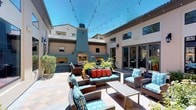This single-story property showcases a generously sized outdoor courtyard that serves as a central entertaining hub. The patio's most commanding feature is a bold teal stucco fireplace structure, architecturally designed with a wide chimney stack, flanked by built-in niche shelving and topped with a broad stone hearth ledge. The fireplace serves as a striking focal point, adding both visual drama and functional warmth to the outdoor space.

The courtyard is finished with neutral-toned stone tile flooring laid in a varied pattern, providing a durable and attractive foundation. Comfortable woven rattan seating — including sofas and armchairs — is arranged around a low coffee table, upholstered in coordinating teal and patterned cushions with vibrant orange accent pillows, creating a lively, inviting atmosphere.

String lights are draped overhead across the open-air space, enhancing the ambiance for evening gatherings. Lush potted plants and greenery add natural warmth throughout the courtyard. The surrounding stucco exterior walls feature large windows and glass doors, seamlessly connecting the interior to this well-appointed outdoor living area under open skies.
