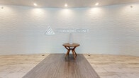 The Paramount lobby in Denver, Colorado presents a sophisticated and contemporary entrance experience that sets an elegant tone for the building. The generously sized single-floor lobby features a striking curved accent wall adorned with a textured wave pattern, prominently displaying The Paramount's logo and serving as a refined focal point upon entry. Light-toned large-format tile flooring flows throughout the space, complemented by warm wood-look flooring details in select areas, creating visual contrast and depth. The entryway showcases a dramatic geometric pendant light fixture in a warm golden hue, drawing the eye upward and casting an inviting glow over the tiled foyer. Nature-inspired wallcoverings featuring a birch tree motif add an organic, artistic element to the walls. A comfortable seating area with sleek, modern black furniture arranged on a rich terracotta-toned area rug provides a welcoming lounge space for residents and guests. Recessed lighting throughout ensures the lobby is well-lit, while large windows near the entry allow natural light to filter in. The overall design blends clean architectural lines with thoughtfully curated décor, creating a polished, upscale atmosphere that reflects the building's commitment to modern, refined living.