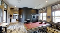 Located at 13195 West Progress Avenue in Littleton, Colorado, this single-floor amenity space offers a well-appointed recreational and social environment designed with modern aesthetics in mind. The space centers around a full-size billiards table set atop a boldly patterned carpet, creating a distinct focal point for leisure and entertainment. A sleek, geometric pendant light fixture hangs above the table, complementing the contemporary design language throughout. A curved dark wood accent wall serves as a dramatic architectural feature, adding depth and texture to the interior while housing a wall-mounted flat-screen television. Adjacent to the billiards area, a small bistro-style seating nook with two chairs and a round table provides an intimate spot for conversation. Large windows along one side of the space invite abundant natural light, offering views of the surrounding greenery and enhancing the open, airy atmosphere. Recessed lighting throughout ensures the space is well-lit at all hours. The open layout allows sightlines into connected areas of the facility, reinforcing the communal and social character of the space. The overall design balances comfort and style, making it a versatile destination for both relaxation and recreation.
