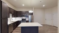 Unit 4308 is a single-floor residence showcasing a contemporary design with a cohesive neutral color palette throughout. The kitchen serves as a central highlight, featuring rich dark espresso cabinetry extending to the ceiling for ample storage, a white subway tile backsplash, light quartz countertops, and a full suite of stainless steel appliances including a refrigerator, range, microwave, and dishwasher. A navy blue center island with a white quartz top provides additional workspace and is accented by a modern pendant light overhead. Recessed lighting further brightens the space. Wood-look plank flooring flows through the kitchen and extends into the open living area, which benefits from natural light provided by large windows fitted with white blinds. The bedroom is generously sized, finished with plush neutral carpet and dual windows that invite natural light. The bathroom features dark vanity cabinetry matching the kitchen, a light quartz countertop, a framed mirror with lighted sconce, and a tub-shower combination. The overall design is clean, modern, and well-coordinated throughout.