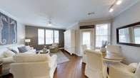 Located at 170 Rebellion Farms Pl in Charleston, South Carolina, this single-story home offers a welcoming and well-appointed interior designed with both comfort and functionality in mind. The open-concept living and dining area serves as the heart of the home, showcasing rich hardwood flooring that flows seamlessly throughout the space. Neutral tones — including soft whites, warm creams, and cool grays — create a cohesive and inviting atmosphere. The living area is anchored by a ceiling fan and flanked by multiple windows that allow generous natural light to pour in, illuminating the space and highlighting its clean, classic finishes. Crown molding adds a polished architectural detail along the ceilings. A striking white armoire provides both a focal point and practical storage, complementing the room's coastal-inspired aesthetic. The dining area features slipcovered chairs gathered around a wood table, evoking a relaxed yet refined sensibility well-suited to the Charleston lifestyle. Overall, this home presents a bright, airy ambiance with thoughtful design choices that balance casual living with timeless style.