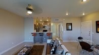 This single-floor residence showcases a thoughtfully designed open-concept layout that seamlessly connects the living and kitchen areas. The living space is anchored by a plush sofa accented with bold patterned throw pillows, a textured area rug, and a ceiling fan with integrated lighting that adds both function and character to the room.

The kitchen is a standout feature, offering warm wood-toned cabinetry, a mosaic tile backsplash, and stainless steel appliances including a refrigerator and microwave. Pendant lighting in rich red tones hangs above the kitchen area, adding a pop of color and warmth. A breakfast bar with a dark wood finish and modern bar stools provides a casual dining option, bridging the kitchen and living areas with an inviting flow.

Recessed lighting throughout ensures the space is well-lit, while neutral-toned walls and light-colored flooring create a clean, airy ambiance. A hallway visible from the living area leads to additional rooms, hinting at a well-organized floor plan. Decorative wall art adds personality to the space. Overall, the residence combines contemporary design elements with practical features, resulting in a cohesive and comfortable living environment.