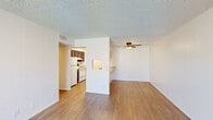 This single-floor unit offers a clean, functional layout with consistent hardwood-style flooring throughout. The open living area is bright and spacious, featuring a ceiling fan with light fixture and a textured ceiling. A half-wall pass-through connects the living space to the kitchen, which is equipped with dark wood cabinetry, white appliances including a range and refrigerator, and a countertop that extends into the living area as a convenient breakfast bar with an integrated sink. The kitchen is accessible through an adjoining hallway, offering both connectivity and defined separation from the main living space. The bedroom is generously sized, featuring the same hardwood-style flooring, white walls, and a window that allows natural light to filter in. The bathroom includes a single-basin vanity with dark wood cabinetry, a stone-tone countertop, a well-lit mirror, and a full-size soaking tub with a shower and curtain rod. Additional storage is accessible from within the bathroom area. Throughout the unit, white walls, clean trim, and neutral tones create a cohesive, move-in-ready aesthetic. The layout is efficient and practical, making good use of the available space on a single level.