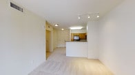 This single-floor space features an open-concept design that seamlessly connects the living area to the kitchen. The living space is finished with plush, light-toned carpeting, lending a comfortable and inviting atmosphere. A track lighting fixture with multiple adjustable heads illuminates the main area, while flush-mount ceiling lights provide consistent brightness throughout the hallway and adjoining spaces.

The kitchen is defined by a raised peninsula counter with a granite-style surface, offering both a functional workspace and a natural divider between the cooking and living areas. Warm medium-toned wood cabinetry lines the kitchen walls, complemented by a built-in appliance visible beneath the upper cabinets. A large fluorescent ceiling fixture ensures the kitchen is well-lit.

A wide hallway leads toward what appears to be additional rooms and a entry door, providing a clear sense of flow throughout the unit. The walls are finished in a neutral cream tone, contributing to the bright and airy feel of the space. Ceiling-mounted HVAC vents are also visible, indicating a central climate control system.