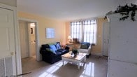 This single-floor space offers a welcoming and well-lit living area characterized by warm yellow-toned walls that create a cozy, cheerful ambiance. The living room is generously sized, accommodating a dark navy sofa accented with colorful throw pillows and a classic white armchair, centered around a charming white-painted wooden coffee table. Natural light flows in abundantly through large windows dressed with plaid patterned shades, brightening the neutral carpeted flooring throughout. Crown molding along the ceiling adds a subtle architectural detail that enhances the room's polished appearance. A framed piece of wall art and a traditional table lamp contribute to the space's homey character. An entry door with a small rug nearby provides convenient direct access. Adjacent to the living area, a glimpse into what appears to be a bathroom or utility space is visible, featuring white cabinetry and bright lighting, suggesting practical and functional amenities nearby. Overall, the space presents a clean, comfortable, and move-in ready environment with a warm, lived-in charm suited for everyday living.