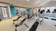 The fitness center at The Lodges On English Station is a thoughtfully designed, single-floor facility that provides residents with a comprehensive workout environment. The space is visually distinguished by bold teal geometric accent walls that contrast with neutral white and gray tones, creating an energetic yet polished atmosphere throughout.

The gym is divided into functional zones that accommodate a variety of training styles. A dedicated cardio area lines one wall and features multiple treadmills and elliptical machines, each equipped with individual monitors. Several windows along this section allow natural light to fill the space and offer outdoor views, enhancing the overall workout experience.

The strength training area is well-stocked with a range of Precor cable machines, multi-functional weight stations, and TAG-branded adjustable benches. A large mirror wall runs along one side of the room, visually expanding the space while allowing users to monitor their form. A separate free weight section provides a full rack of dumbbells in varying sizes, paired with additional bench options.

Decorative pendant light fixtures hang from the vaulted ceiling alongside recessed lighting, contributing to a warm and welcoming ambiance. Durable, light-toned flooring covers the entire facility, offering comfort and easy maintenance. A glass-panel interior window connects two sections of the gym, maintaining an open, airy feel throughout.