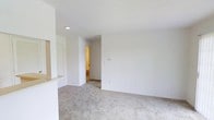 This single-floor space presents a straightforward, functional layout defined by clean lines and a neutral color palette. The main living area is generously proportioned, featuring wall-to-wall plush carpeting in a light beige tone that adds warmth underfoot and contributes to the room's understated, comfortable ambiance. Bright white walls throughout create a sense of openness and provide a versatile backdrop adaptable to a variety of interior styles.

Recessed lighting in the ceiling offers focused illumination, complementing the overall clean aesthetic. A partial wall or breakfast bar divides the living area from an adjacent space, hinting at a connected kitchen or dining area and adding a subtle architectural element to the otherwise open floor plan. Multiple electrical outlets are positioned along the walls, offering practical flexibility for furniture arrangement.

A hallway visible from the main living area leads to additional rooms, suggesting a well-organized layout that maintains separation between living and private spaces. The warmly lit corridor adds depth to the interior. Overall, the space maintains a simple, move-in-ready condition with consistent finishes throughout.