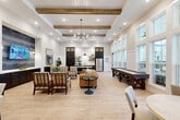Located in Dalton, GA, this thoughtfully designed community living room blends modern farmhouse aesthetics with a warm, inviting ambiance. The space is defined by soaring tray ceilings adorned with rustic exposed wood beams, shiplap detailing, and a statement sputnik chandelier that anchors the room with elegance. Light hardwood flooring stretches throughout, complementing the bright white walls and creating a cohesive, airy feel. A wall of floor-to-ceiling windows along one side bathes the interior in natural light while offering views of the surrounding outdoor terrace and community. The living area features a well-curated arrangement of plaid armchairs, a leather sofa, and accent tables, creating an ideal gathering space. Adjacent to the seating area, a full-length shuffleboard table provides entertainment along the window wall. A built-in kitchenette with dark cabinetry, glass-front upper cabinets, open shelving, a stainless steel refrigerator, and a coffee station serves residents with ease. A long communal dining table with black spindle chairs provides a dedicated space for shared meals or gatherings. Large-scale artwork and a wall-mounted TV round out the space, adding personality and function to this well-appointed community retreat.