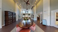 This thoughtfully designed single-floor community clubhouse offers an impressive and inviting atmosphere from the moment you enter. The grand foyer greets visitors with soaring double-height ceilings, rich dark hardwood flooring, and an abundance of natural light pouring through oversized windows adorned with clean white blinds. A central round table with upholstered ottomans serves as a welcoming focal point near the entrance.

The expansive main lounge is anchored by a striking floor-to-ceiling stacked stone fireplace, framed by elegant accent chairs and complemented by lush indoor trees that add a natural, organic element to the space. Multiple seating areas are arranged throughout, including a generous sectional sofa and coordinating furniture, providing comfortable spaces for gathering and relaxation.

A well-appointed kitchenette features rich dark cabinetry, granite countertops, a decorative tile backsplash, a built-in wine rack, and full-size appliances — ideal for hosting events and catering needs. A separate dining room accommodates group gatherings beneath a modern rectangular chandelier, with a dramatic large-scale mural as a bold artistic backdrop. A wall-mounted display screen and additional corridor access round out the functional amenities of this well-curated community space.