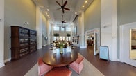 This thoughtfully designed single-floor community clubhouse offers an impressive and inviting atmosphere from the moment you enter. The grand foyer greets visitors with soaring double-height ceilings, rich dark hardwood flooring, and an abundance of natural light pouring through oversized windows adorned with clean white blinds. A central round table with upholstered ottomans serves as a welcoming focal point near the entrance.

The expansive main lounge is anchored by a striking floor-to-ceiling stacked stone fireplace, framed by elegant accent chairs and complemented by lush indoor trees that add a natural, organic element to the space. Multiple seating areas are arranged throughout, including a generous sectional sofa and coordinating furniture, providing comfortable spaces for gathering and relaxation.

A well-appointed kitchenette features rich dark cabinetry, granite countertops, a decorative tile backsplash, a built-in wine rack, and full-size appliances — ideal for hosting events and catering needs. A separate dining room accommodates group gatherings beneath a modern rectangular chandelier, with a dramatic large-scale mural as a bold artistic backdrop. A wall-mounted display screen and additional corridor access round out the functional amenities of this well-curated community space.