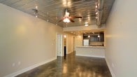 This single-floor space blends industrial character with modern functionality, creating a distinctive and open atmosphere throughout. The living area commands attention with its polished concrete floors, which reflect light and lend a sleek, urban feel to the interior. Overhead, a corrugated metal ceiling adds a bold industrial aesthetic, complemented by exposed round ductwork that runs the length of the space. A ceiling fan with integrated lighting anchors the main living area, while a rail track lighting system provides flexible, directional illumination along the ceiling. The open layout flows seamlessly toward the kitchen, which is defined by a white breakfast bar counter that serves as a natural dividing element between the living and culinary spaces. Dark cabinetry lines the kitchen walls, offering generous storage, while pendant lighting descends above the bar area, adding warmth and task-focused light. Recessed ceiling lights in the kitchen zone ensure the workspace is well-lit. A white refrigerator and integrated appliances complete the kitchen's functional design. Toward the rear of the space, doorways lead to additional rooms, including what appears to be a bathroom and further private areas. Neutral-toned walls throughout keep the overall ambiance bright and adaptable, allowing the industrial architectural details to remain the defining feature of this well-designed space.