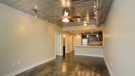 This single-floor space blends industrial character with modern functionality, creating a distinctive and open atmosphere throughout. The living area commands attention with its polished concrete floors, which reflect light and lend a sleek, urban feel to the interior. Overhead, a corrugated metal ceiling adds a bold industrial aesthetic, complemented by exposed round ductwork that runs the length of the space. A ceiling fan with integrated lighting anchors the main living area, while a rail track lighting system provides flexible, directional illumination along the ceiling. The open layout flows seamlessly toward the kitchen, which is defined by a white breakfast bar counter that serves as a natural dividing element between the living and culinary spaces. Dark cabinetry lines the kitchen walls, offering generous storage, while pendant lighting descends above the bar area, adding warmth and task-focused light. Recessed ceiling lights in the kitchen zone ensure the workspace is well-lit. A white refrigerator and integrated appliances complete the kitchen's functional design. Toward the rear of the space, doorways lead to additional rooms, including what appears to be a bathroom and further private areas. Neutral-toned walls throughout keep the overall ambiance bright and adaptable, allowing the industrial architectural details to remain the defining feature of this well-designed space.