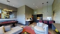 Located at 1-3 Mill River Lane in Ardsley, NY 10502, this single-floor space is a thoughtfully designed amenity lounge that combines functionality with contemporary style. The space features soaring vaulted ceilings that create an open, airy atmosphere, complemented by recessed lighting and sleek pendant fixtures that add a modern touch.

The layout is divided into distinct zones for relaxation and entertainment. A comfortable seating area anchors the room, furnished with plush sofas and accent chairs arranged around a central coffee table, with natural wood stump side tables adding an organic, earthy element. A vibrant red area rug grounds the lounge area and introduces a bold pop of color against the neutral gray wall tones.

A billiards table occupies one side of the space, making it an ideal spot for recreation and social gatherings. On the opposite end, a well-equipped kitchenette provides bar-height seating, upper and lower cabinetry, a microwave, and countertop appliances, offering a convenient space for casual dining or entertaining. The flooring transitions between tile and wood-toned surfaces, adding visual texture and definition throughout. Overall, the space delivers a refined, inviting environment suited for both leisure and community use.