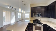 Plan H at 5945 Linda Vista offers a thoughtfully designed single-floor layout that emphasizes open living. The kitchen is a standout feature, outfitted with rich dark cabinetry, light-toned countertops, and stainless steel appliances including a gas range and built-in microwave. A large kitchen island extends the counter space and serves as a natural dividing point between the kitchen and the adjoining living area. Three sleek pendant lights hang above, adding a contemporary visual accent while providing focused illumination over the workspace.

The open-concept design allows the kitchen to flow directly into the living space, creating a cohesive and spacious feel throughout. The living area benefits from multiple windows that invite generous natural light, contributing to a bright and airy ambiance. Track lighting along the ceiling supplements the natural light and enhances the modern aesthetic. The neutral color palette — soft walls paired with light flooring — lends a clean, welcoming atmosphere throughout the space. Vertical blinds on the windows offer privacy while maintaining the room's light-filled character.