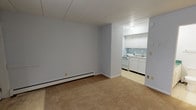 This single-floor unit offers a straightforward, functional layout designed for comfortable everyday living. The main living area is finished with wall-to-wall carpet in a neutral tan tone and painted in soft, light gray hues, creating a calm and understated atmosphere. A baseboard heating system runs along the exterior wall, and a large window fitted with vertical blinds allows natural light to fill the space while providing privacy. A door off the main room provides direct access to the exterior.

The kitchen, accessible directly from the living area through an open passage, features white cabinetry, a white gas range, a sink, and a light blue tile backsplash that adds a subtle touch of color. Overhead lighting keeps the kitchen bright and practical.

The bedroom is carpeted and includes a built-in sliding-door closet with shelving, offering organized storage. The neutral walls and consistent flooring tie the room seamlessly into the rest of the unit.

The bathroom is outfitted with a combination tub and shower with a curtain rod, a toilet, and a vanity topped with a distinctive green marbled countertop that serves as a visual accent. A towel bar and baseboard heating complete the space. Overall, the unit presents a clean, no-frills interior ready for personalization.