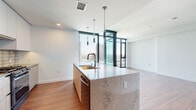 Located in the heart of downtown San Diego, this contemporary high-rise unit at 675 9th Ave offers a refined urban living experience defined by clean lines, premium materials, and sweeping city views. The open-concept layout is anchored by warm hardwood-style flooring that flows seamlessly throughout the space, complemented by crisp white walls and recessed lighting.

The kitchen is a standout feature, showcasing a large central island topped with stone-veined quartz countertops and an integrated sink with a matte black faucet. Sleek flat-front cabinetry in a light wood tone pairs with white upper cabinets, a textured tile backsplash, and stainless steel appliances including a French door refrigerator, gas range, and dishwasher. Cylindrical pendant lights above the island add a modern, industrial touch.

Floor-to-ceiling windows and glass doors flood the living area with natural light while framing panoramic views of the San Diego skyline and surrounding cityscape, creating a dramatic sense of openness and elevation. The bedroom continues this airy aesthetic with expansive window walls offering additional city views.

The bathroom is appointed with a floating vanity in light wood with quartz countertops, a matte black fixture, an under-mount sink, and a backlit mirror that lends a soft, ambient glow. The overall design is cohesive and contemporary, offering a polished urban retreat in one of San Diego's most vibrant neighborhoods.