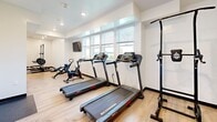 Located on Laurel Canyon Boulevard in Los Angeles, this fitness center offers a thoughtfully designed workout space that accommodates a comprehensive range of exercise needs. The room features light hardwood-style flooring throughout, crisp white walls, and recessed lighting that creates a clean, energizing atmosphere. Multiple windows fitted with horizontal blinds allow natural light to fill the space while maintaining privacy, contributing to an open and welcoming environment.

The layout is divided into functional zones. The cardio area is well-appointed with multiple Schwinn treadmills and a stationary cycling bike, all positioned to take advantage of the natural light from the surrounding windows. A wall-mounted flat-screen television is also present, enhancing the cardio experience. The strength training zone features a multi-function cable machine, a squat rack with an Olympic barbell and weight plates, an adjustable workout bench, and a power tower for bodyweight exercises. Rubber matting is placed beneath the free weight equipment for added safety and floor protection.

The ceiling height provides a comfortable sense of openness throughout the space. With its modern, minimalist design and diverse selection of equipment, this fitness center is well-suited to support a variety of training routines.