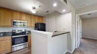 Located in Saint Petersburg, FL, this one-bedroom, two-bathroom residence offers a well-designed single-floor layout that makes efficient use of its space. The kitchen is a functional highlight, featuring warm honey-toned cabinetry, dark granite countertops, wood-look flooring, and a full suite of appliances including a stainless steel range, built-in microwave, dishwasher, and black refrigerator. A peninsula with a granite countertop provides additional prep space and a natural transition into the living area. The living room is bright and welcoming, with large windows and a glass-paned door that flood the carpeted space with natural light and offer views of the surrounding greenery. The bedroom is a comfortable retreat with plush carpeting and access to a dedicated walk-in closet equipped with dual wire shelving systems for ample storage. Both bathrooms are appointed with warm wood vanities, granite countertops, and large mirrors. One bathroom features a full tub-shower combination with clean white tile surround and wood-look flooring. Throughout the residence, a textured ceiling, neutral wall tones, and consistent finishes contribute to a cohesive and livable atmosphere.