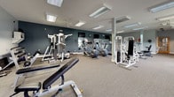 This fitness center offers a spacious, single-floor layout designed to accommodate a wide range of workout needs. The facility is divided into distinct functional zones, with cardio equipment positioned near large windows that allow natural light to fill the space, creating an open and energizing atmosphere. Treadmills, upright and recumbent stationary bikes, and additional cardio machines are arranged to provide ample room for movement between stations.

The strength-training area features multiple cable and weight machines, including Paramount-branded multi-station units, along with adjustable benches and a dedicated free-weight section equipped with a dumbbell rack. Full-length mirrors line one wall, enhancing the sense of depth while allowing users to monitor their form during exercises. A stability ball and floor mat area provide space for stretching and functional training.

The interior is finished with neutral gray walls accented by a deep teal feature wall, carpeted flooring throughout, and recessed and panel lighting supplemented by ceiling fans. A wood-framed glass entry door provides a welcoming entrance, and wall-mounted televisions offer entertainment during workouts. The overall ambiance is clean, functional, and well-maintained.