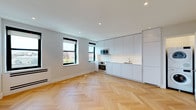 This thoughtfully designed single-floor space combines contemporary aesthetics with functional living. The standout feature throughout is the striking herringbone-patterned hardwood flooring in warm oak tones, which lends a sophisticated character to every room.

The open-concept living area is bright and airy, benefiting from multiple black-framed windows that frame views of the surrounding cityscape and invite generous natural light. Recessed ceiling lighting complements the natural light, ensuring the space is well-illuminated throughout. Clean white walls and smooth ceilings contribute to a minimalist, modern atmosphere.

The kitchen is seamlessly integrated into the living space and features floor-to-ceiling flat-front cabinetry in a soft light grey finish. Stainless steel appliances — including a built-in oven, microwave, and cooktop — provide a polished, professional quality. The white countertops and backsplash maintain a cohesive, understated palette.

The bathroom offers a refined finish with taupe hexagonal mosaic floor tiles, crisp white subway wall tiles, a full soaking tub with an overhead shower, and classic white fixtures. The overall design balances clean lines with tactile detail.

Throughout the space, the consistent use of neutral tones, quality materials, and modern fixtures creates a cohesive, elegant environment suited for contemporary living.