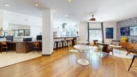This well-appointed single-floor community space blends contemporary design with a welcoming, social atmosphere. Warm hardwood flooring flows throughout, connecting a series of distinct yet open functional zones. The main lounge area features mid-century modern seating arrangements, including leather accent chairs, a round pedestal coffee table, and transparent acrylic dining chairs, all set beneath a stylish ceiling fan and recessed lighting. Large windows dressed with floor-length drapes invite abundant natural light and offer views of the surrounding greenery and outdoor areas. A bar-height counter with black stools serves as a natural dividing element between the lounge and a well-equipped kitchenette. The kitchenette features rich walnut cabinetry, white quartz countertops, a stainless steel sink, a dishwasher, a microwave, and open floating shelves for display. A dedicated workstation area with desktop computers provides a functional space for focused tasks. A bold, navy blue accent room offers a cozy nook with a built-in bench seat, teal cushions, large windows, and vibrant abstract artwork on the walls, creating an intimate retreat within the larger space. Throughout, colorful art pieces and thoughtful décor accents contribute to an energetic yet comfortable community environment.