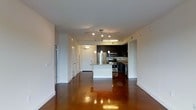 This single-floor space showcases a contemporary open-concept design that seamlessly connects the living area to the kitchen. The most striking feature is the richly toned, high-gloss polished concrete flooring that spans the entire space, reflecting light and adding a warm, industrial-chic aesthetic throughout.

The kitchen is positioned centrally and is anchored by a white island/breakfast bar that provides both workspace and casual seating. Dark espresso cabinetry contrasts sharply with the lighter walls, while stainless steel appliances — including a refrigerator, range, and microwave — provide a sleek, modern finish. The countertops appear to feature a stone or granite surface. Pendant lighting hangs above the island, complementing the track lighting system installed in the main living area, which ensures thorough illumination across the open floor plan.

The living area is spacious and bright, with white walls and a clean, minimalist design. A hallway visible toward the rear leads to additional rooms, lined with several doors suggesting multiple private spaces. The overall ambiance is modern, clean, and versatile.